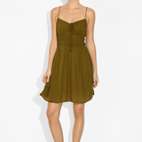 A woman modeling a short olive green spaghetti-strap dress with a tie-front detail and a gathered elastic waist, standing against a plain white studio background.