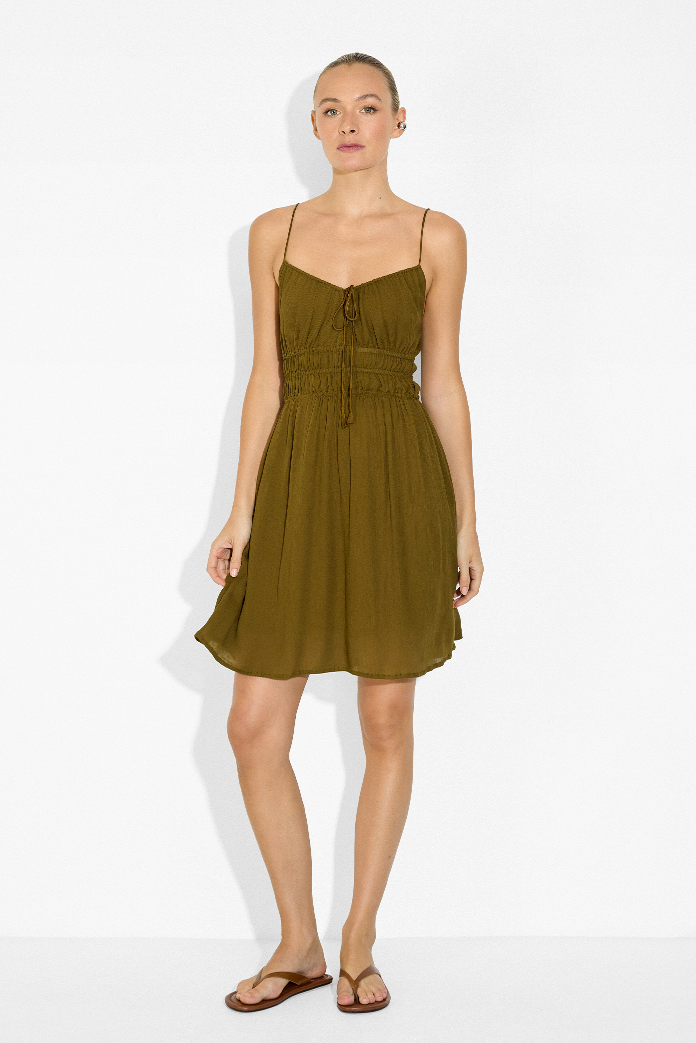 A woman modeling a short olive green spaghetti-strap dress with a tie-front detail and a gathered elastic waist, standing against a plain white studio background.