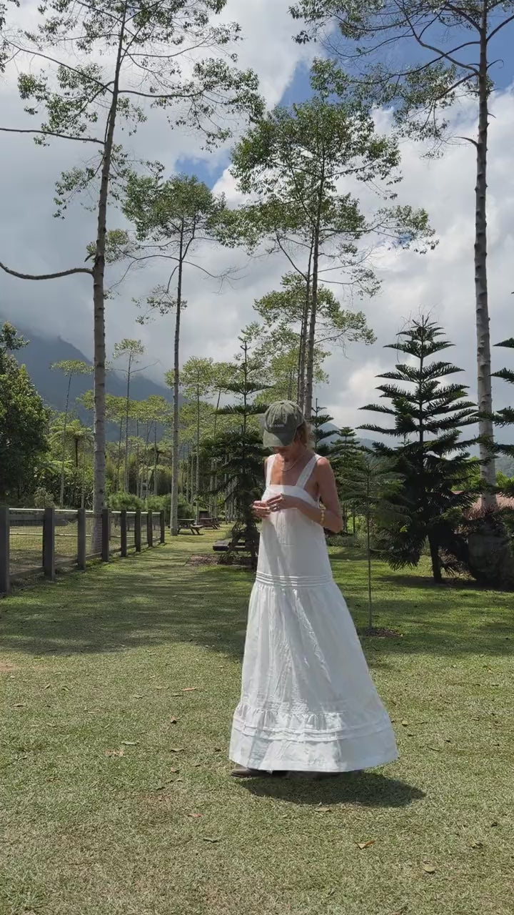 Model walking in the white cotton Aurora Maxi Dress on a lawn lined with pine trees
